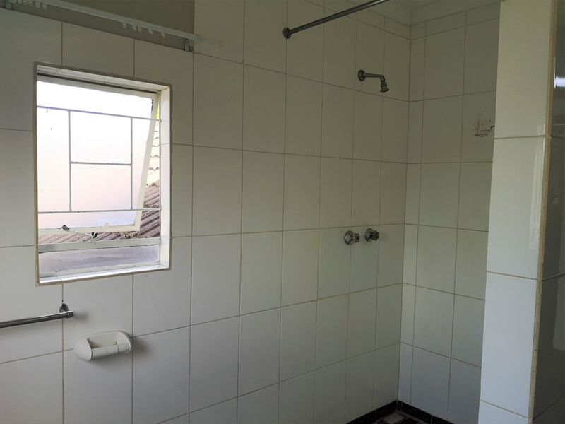 A functional bathroom shower area featuring white wall tiles, a top-hung window with security bars, and basic shower fixtures.