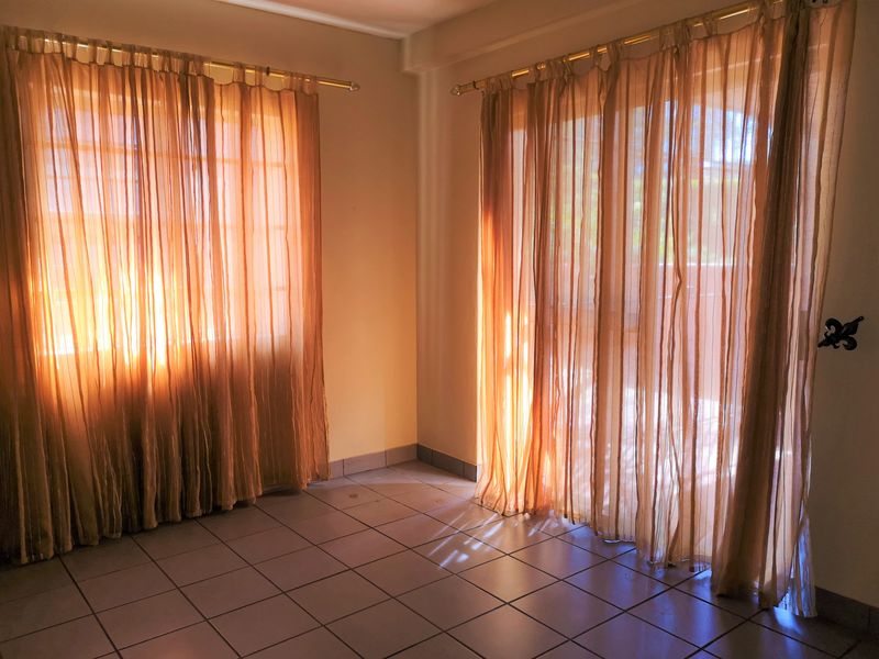 An empty living room featuring tiled flooring and large windows with natural light, providing access to an outdoor area.