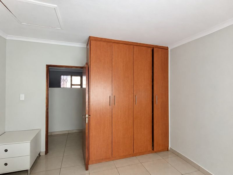 A clean, empty bedroom featuring neutral tiled flooring, built-in wooden cupboards, and a simple, functional layout with access to an adjacent room.