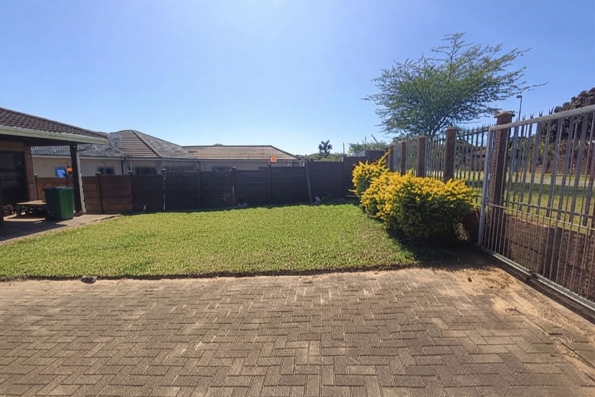 A private rear garden area featuring a paved driveway, a well-maintained lawn, timber boundary fencing, and a secure metal gate.