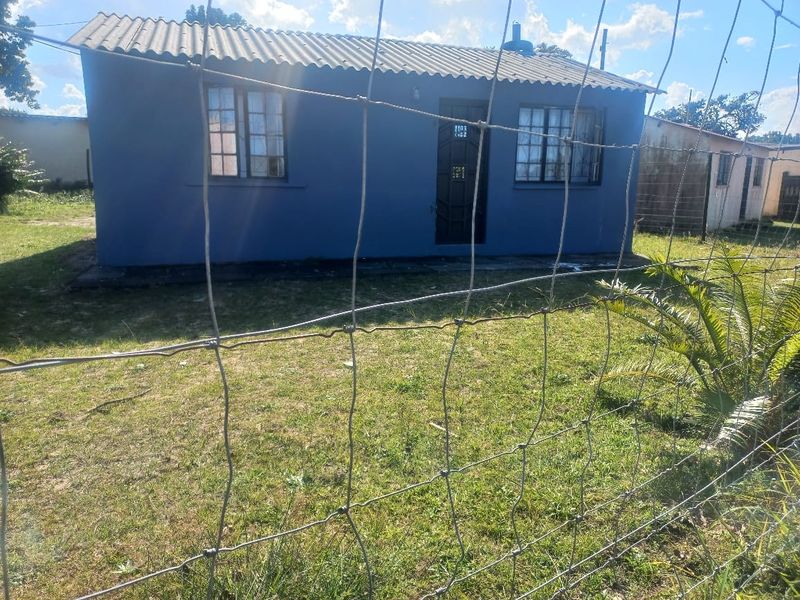 A modest single-story detached house featuring a blue painted exterior, corrugated roof sheeting, and security-barred windows, set within a fenced yard.