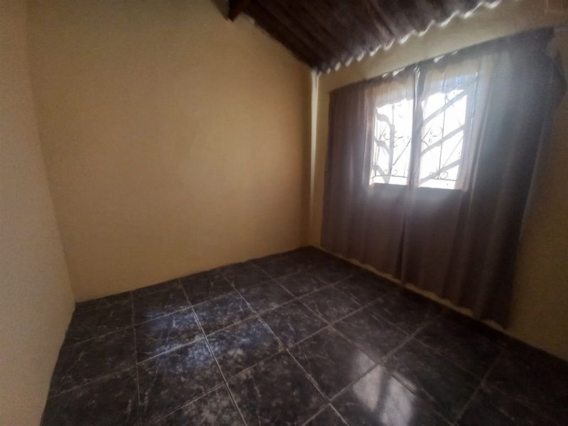 A simple, empty bedroom featuring dark tiled flooring, a window with security bars, and an exposed ceiling structure.