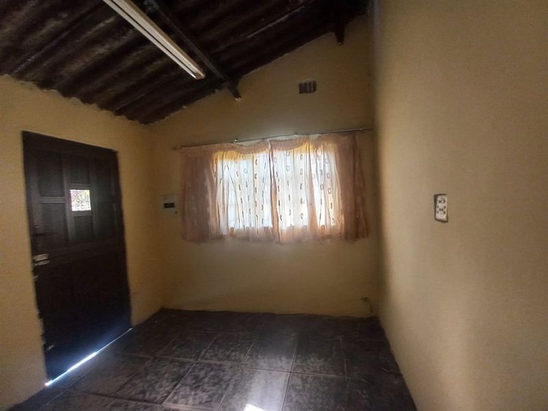 A simple, empty bedroom featuring tiled flooring, a single window with curtains, and an exposed rafter ceiling.