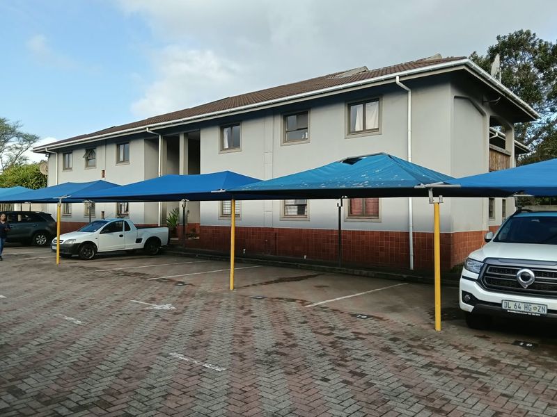 Exterior view of an apartment building featuring a paved parking area with blue shade cloth carports and a tiled roof.