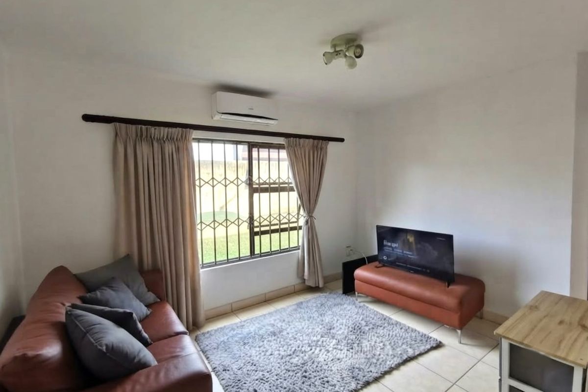 A functional living room featuring tiled flooring, a wall-mounted air conditioning unit, and security bars on the window.