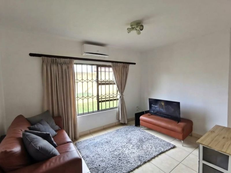 A functional living room featuring tiled flooring, a wall-mounted air conditioning unit, and security bars on the window.