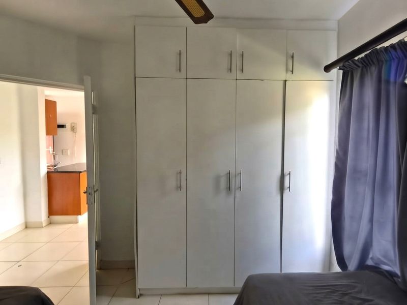 A bedroom featuring white built-in wardrobes, tiled flooring, and a ceiling fan, with a view into an adjacent kitchen area.