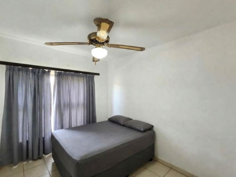 A simple bedroom featuring tiled flooring and a ceiling fan, providing a functional space with neutral walls and window treatments.
