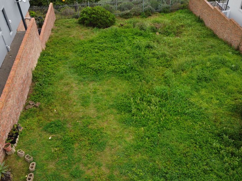 An elevated view of a vacant residential plot enclosed by brick boundary walls and metal fencing, featuring an overgrown grass surface.