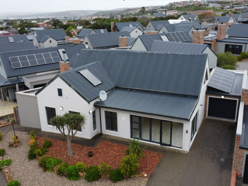 Aerial view of a modern residential property featuring a corrugated metal roof with solar installations, paved driveway, and double garage in a suburban estate.