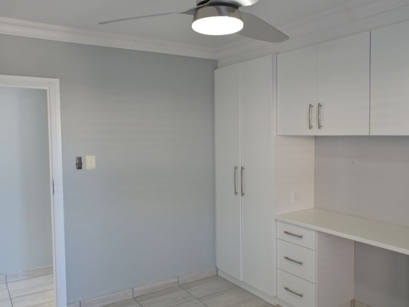 A clean, modern home office featuring built-in white cabinetry, a matching desk, tiled flooring, and a contemporary ceiling fan.