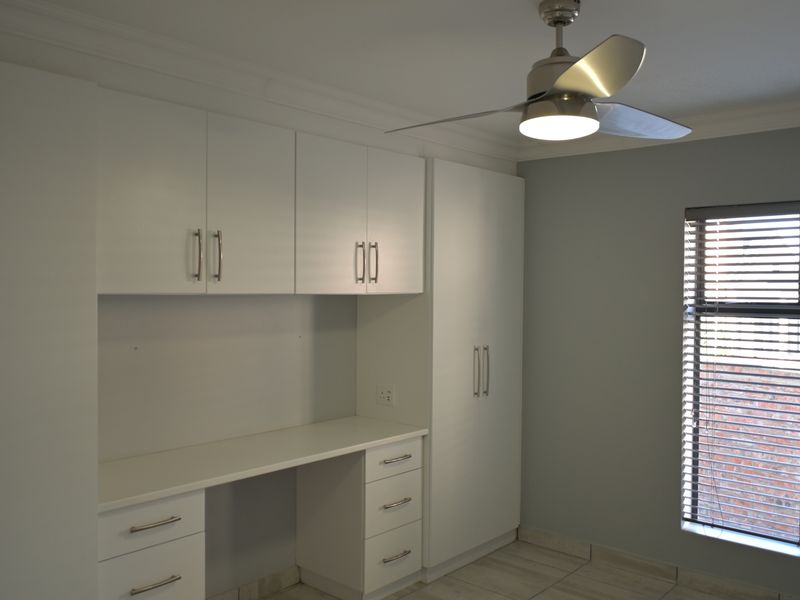 A clean, modern bedroom featuring extensive built-in cabinetry, a dedicated desk area, ceiling fan, and neutral tiled flooring with venetian blinds.