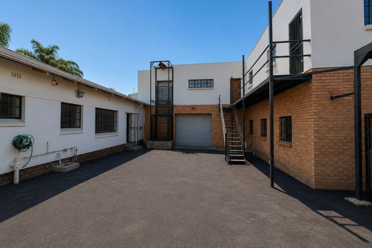 Rear exterior view of the industrial property featuring an asphalt courtyard, roller shutter door access, and an external metal staircase leading to an upper level.