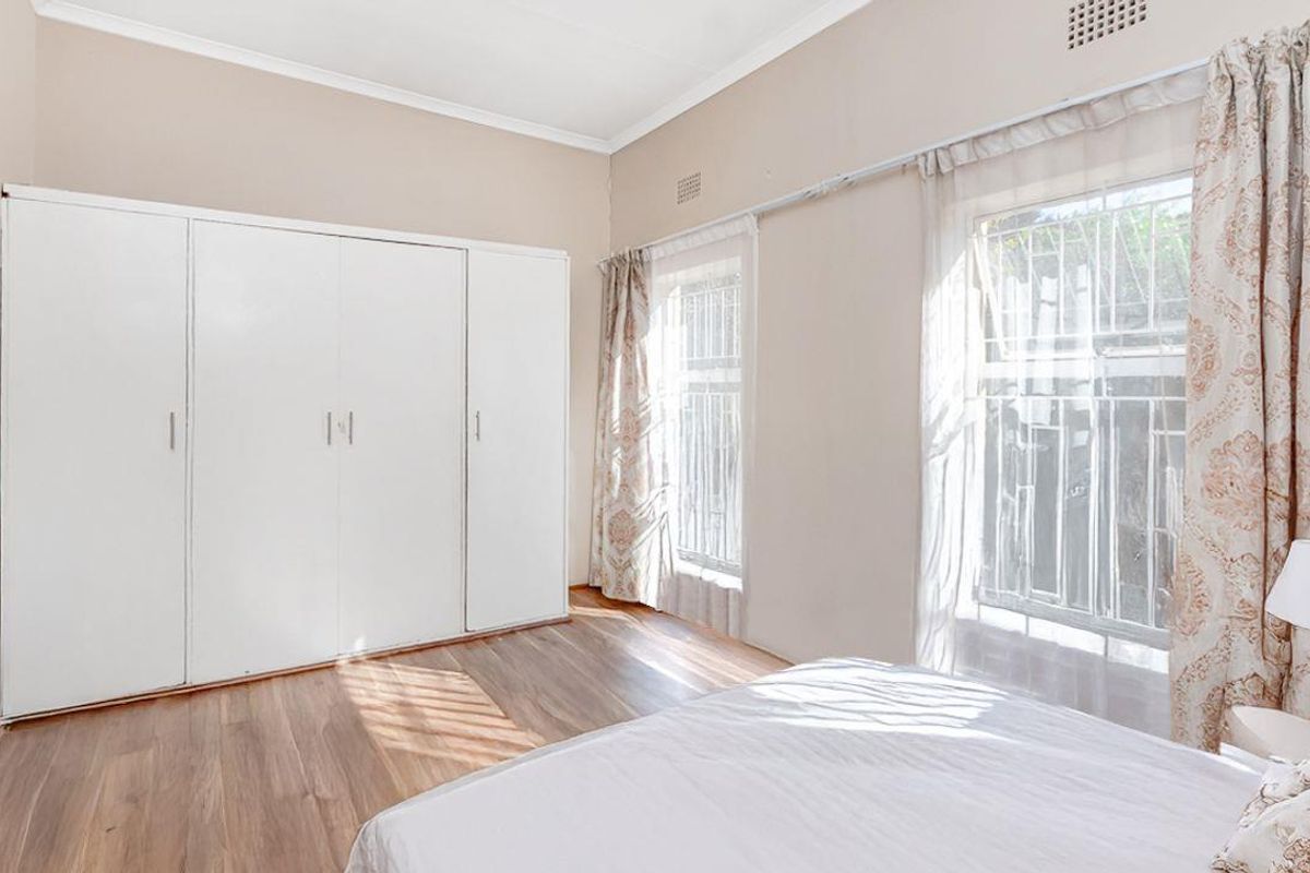 A bright bedroom featuring wood-look flooring, built-in wardrobes, and large windows with security bars, offering a clean and functional living space.