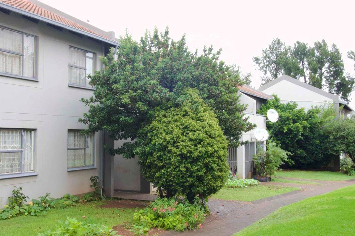 Exterior view of a multi-story apartment complex featuring paved walkways, landscaped garden areas, and established trees surrounding the building facade.