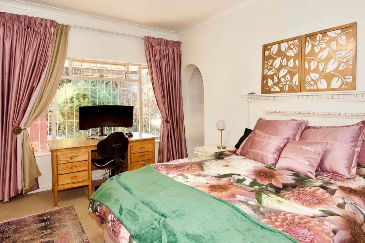 A well-lit bedroom featuring a large window, neutral carpeting, and an arched doorway leading to an adjacent space.
