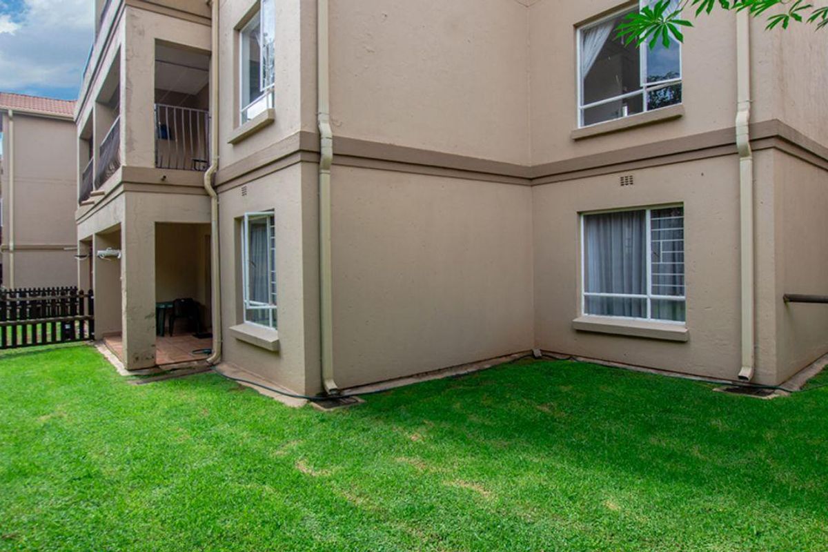 Exterior view of a townhouse complex featuring a ground-floor patio, landscaped lawn, and multi-story building facade with aluminum-framed windows.