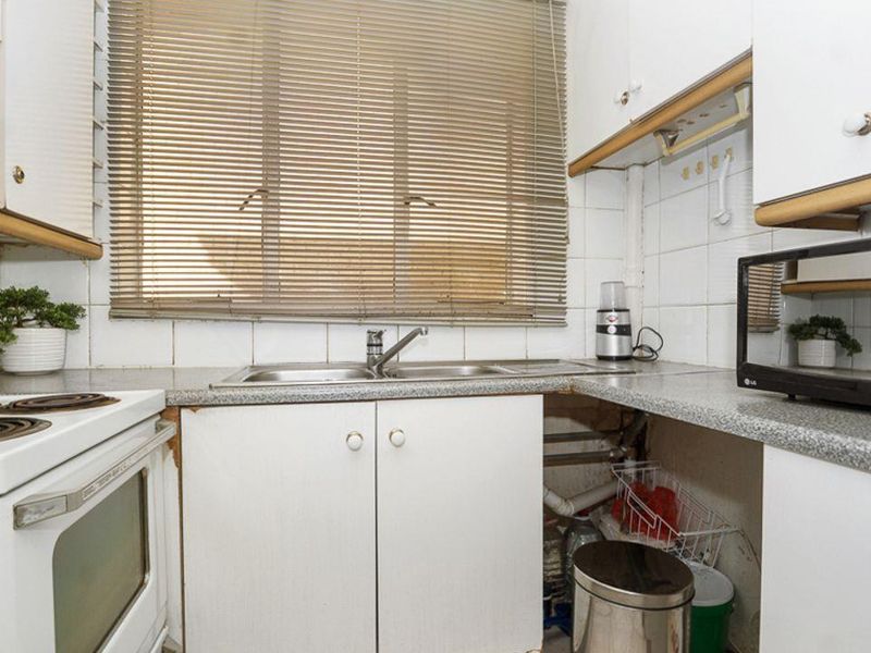 A compact kitchen featuring white cabinetry, laminate countertops, a stainless steel sink, and an electric stove, currently showing signs of wear and exposed plumbing.