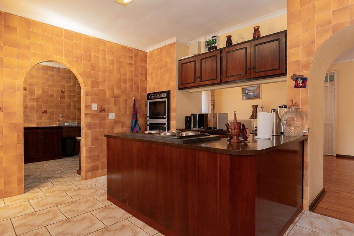 A dated kitchen featuring a central island, built-in oven, hob, and extensive wall tiling, with an open-plan layout connecting to adjacent living spaces.