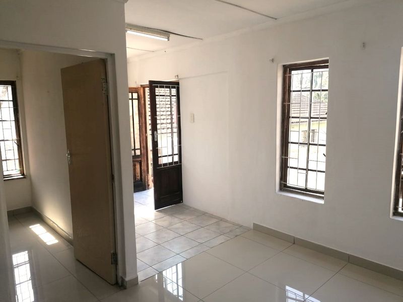 An empty living area featuring tiled flooring, security bars on windows, and direct access to an adjacent room through a timber door frame.