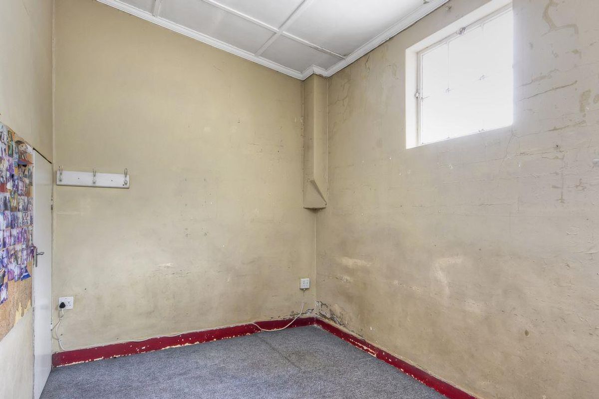 A small, vacant room featuring a high ceiling, a single window, and worn wall and floor finishes in need of renovation.