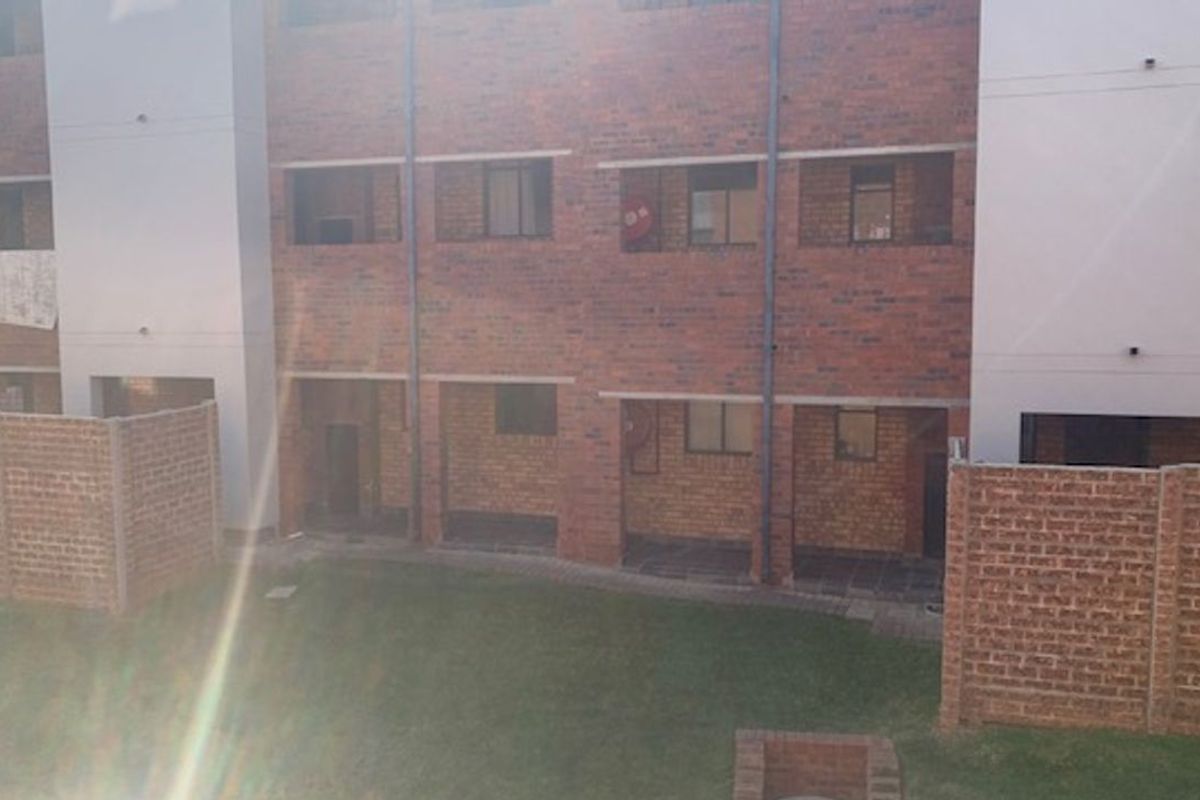 A view of the rear exterior of a multi-story apartment building featuring exposed brickwork, ground-level patio areas, and a shared lawn.