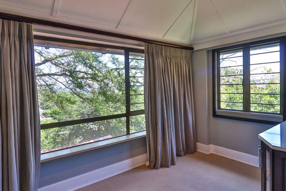 A bright bedroom featuring large windows with security bars, vaulted ceilings, and neutral carpeting, offering pleasant views of the surrounding greenery.