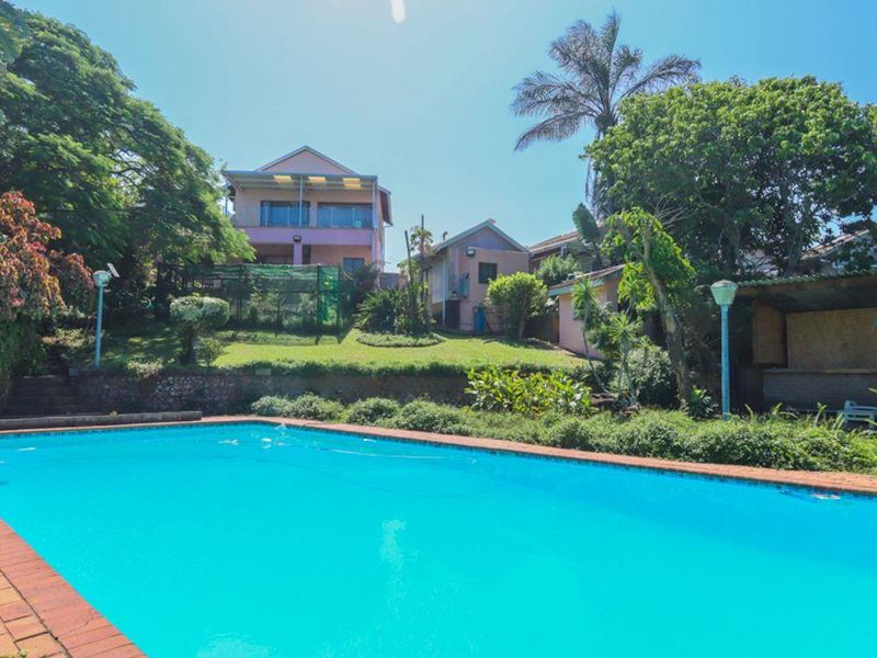 Spacious outdoor pool area featuring a large swimming pool with brick coping, set against a landscaped garden and multi-level residential structures.