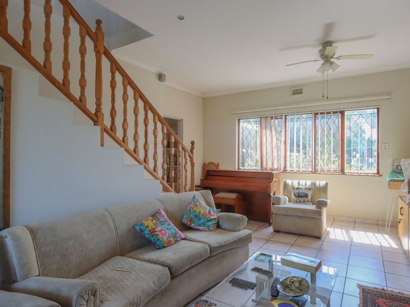 A living room featuring tiled flooring, a wooden staircase with a balustrade, and large windows with security bars, offering a functional and well-lit space.