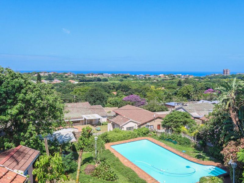 Elevated aerial view overlooking a residential neighborhood with a private swimming pool, lush garden, and distant ocean horizon.