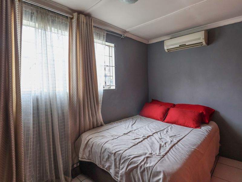 A functional bedroom featuring tiled flooring, a wall-mounted split-system air conditioner, and windows fitted with security bars.