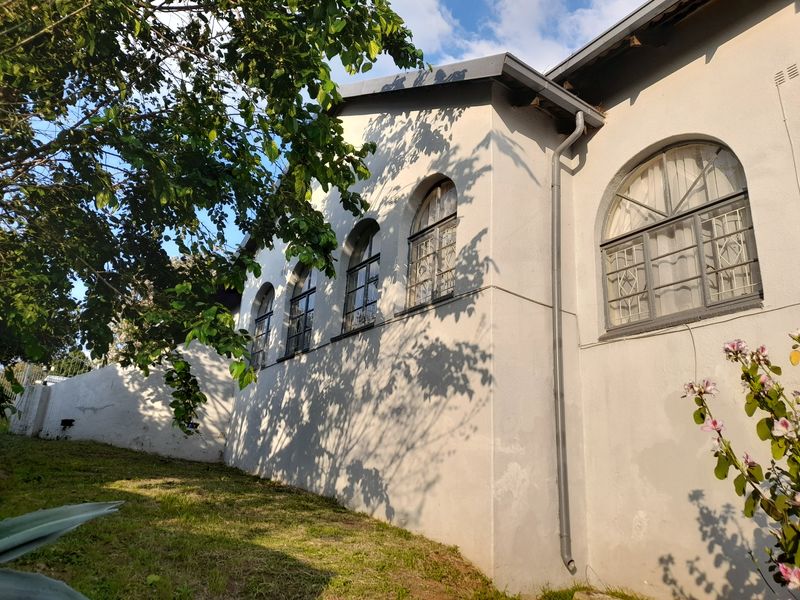 A view of the rear exterior wall featuring arched windows with security bars and a downpipe, set against a garden area.