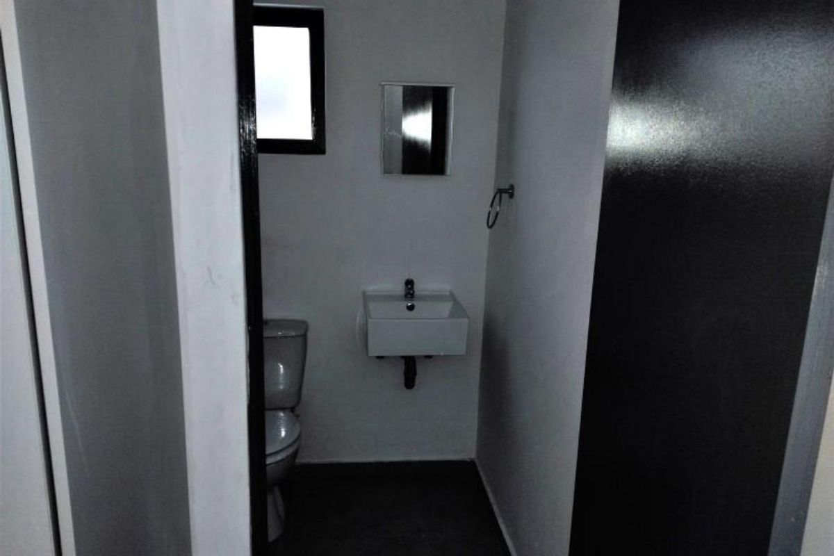 A compact, minimalist bathroom featuring a wall-mounted basin, toilet, and small mirror, finished with neutral walls and dark flooring.