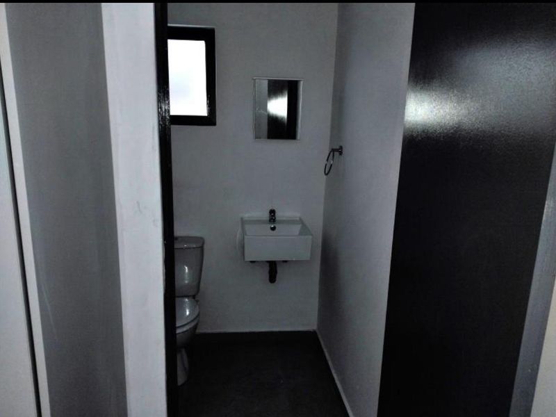 A compact, minimalist bathroom featuring a wall-mounted basin, toilet, and small mirror, finished with neutral walls and dark flooring.