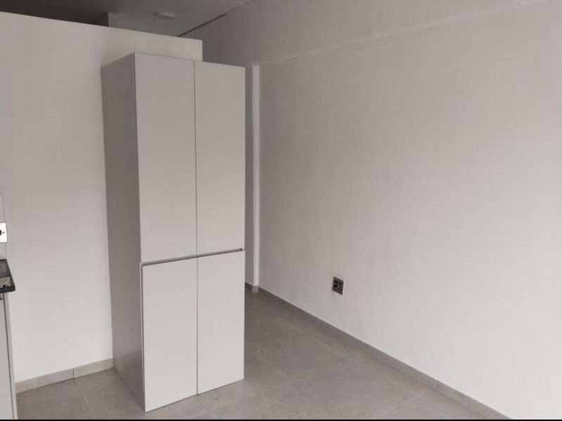 A minimalist kitchen area featuring clean white built-in cabinetry and neutral tiled flooring in a bright, open-plan apartment setting.
