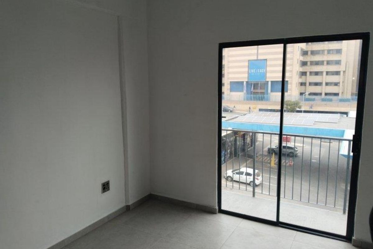 An empty bedroom featuring neutral tiled flooring and a sliding glass door that opens onto a balcony with an urban street view.