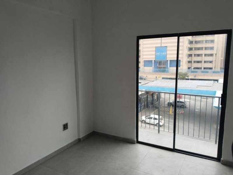 An empty bedroom featuring neutral tiled flooring and a sliding glass door that opens onto a balcony with an urban street view.