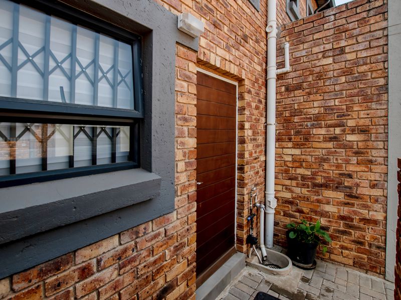 A side exterior view of the townhouse featuring face brick walls, a secure entrance door, and functional external plumbing fixtures.