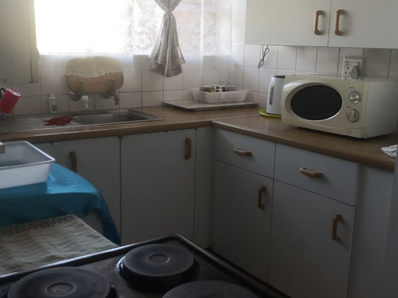A compact, dated kitchen featuring laminate countertops, built-in cabinetry, a stainless steel sink, and an electric stove with a tiled backsplash.