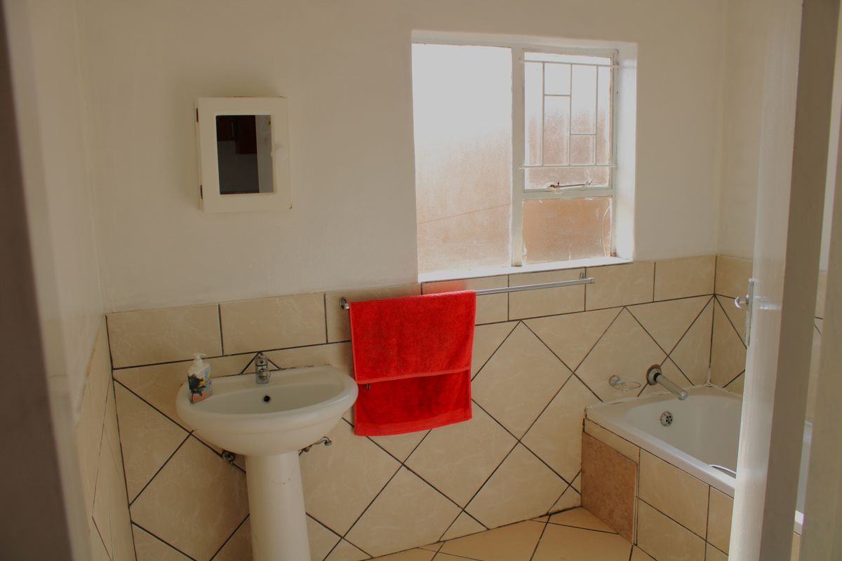A functional bathroom featuring a pedestal basin, bathtub with tiled surround, and a window with security bars.