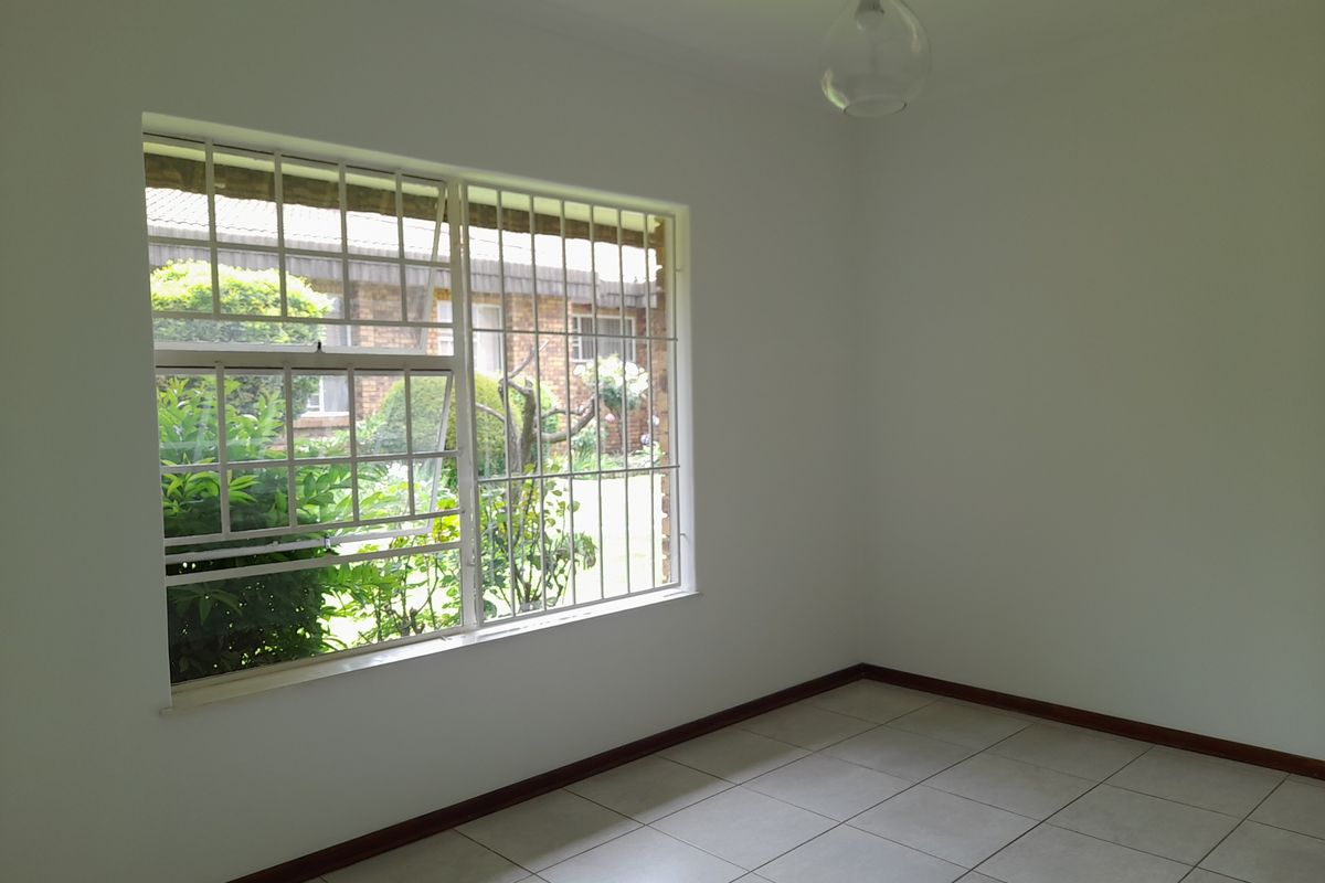 A bright, empty bedroom featuring neutral tiled flooring, white walls, and a large window fitted with security bars overlooking the garden.