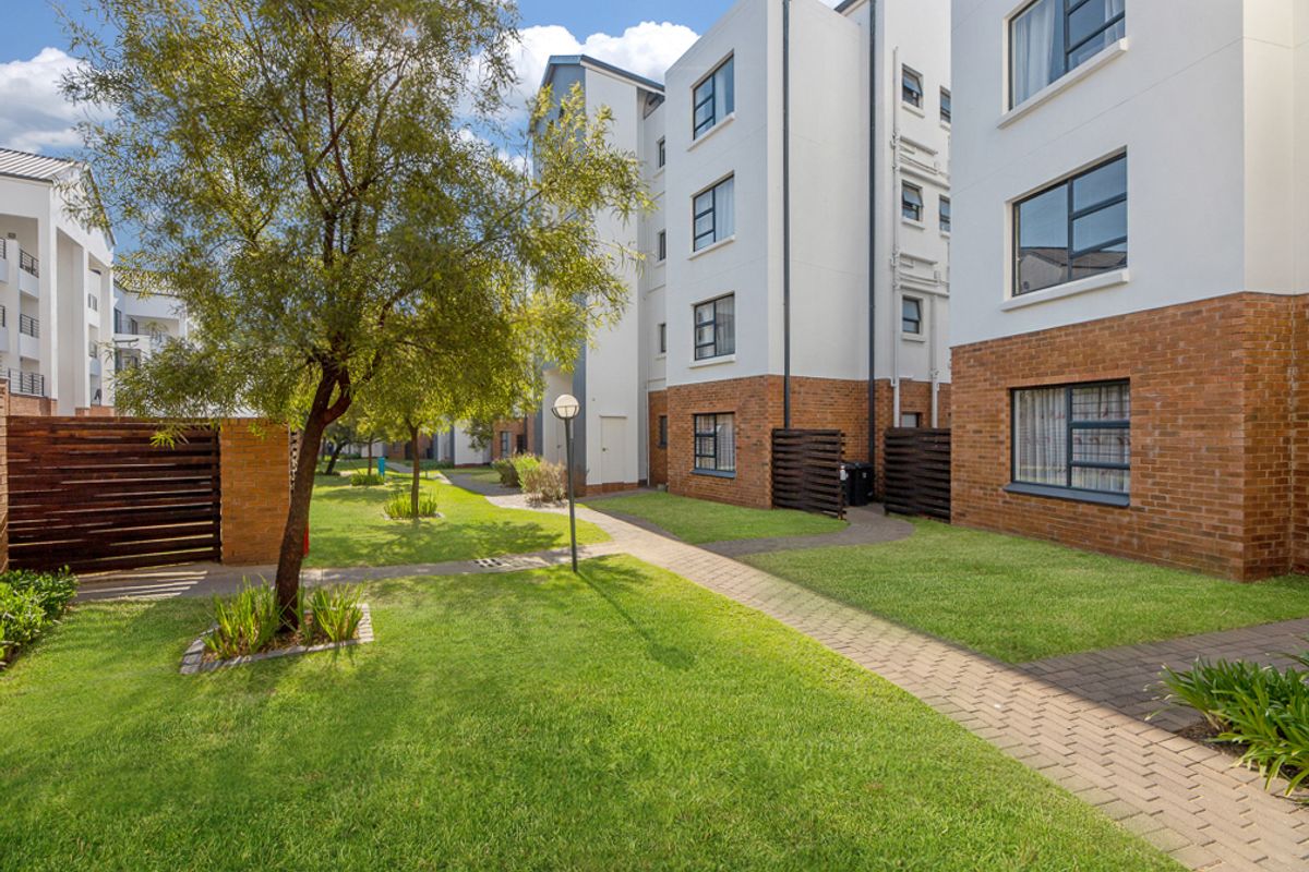 A well-maintained communal garden area featuring paved walkways, manicured lawns, and established trees between modern apartment blocks with brick and plaster facades.