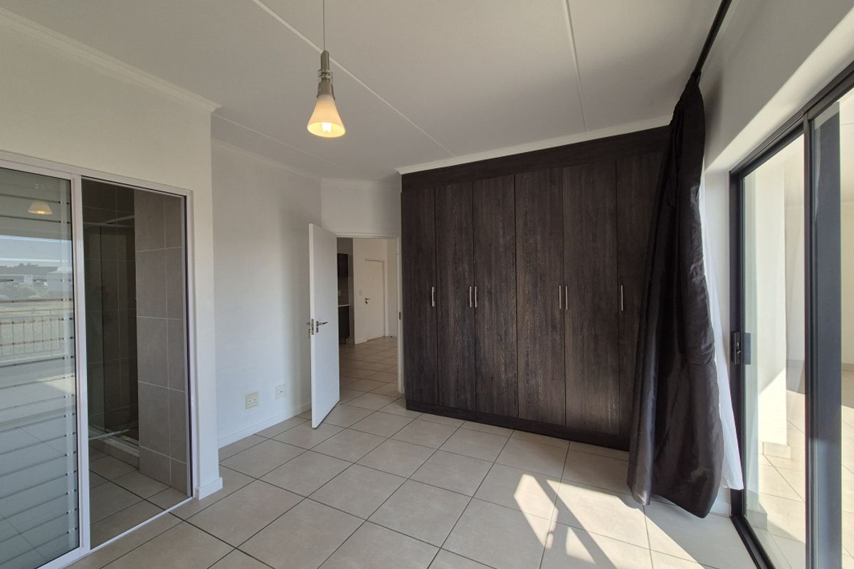 A bright bedroom featuring built-in dark wood cabinetry, tiled flooring, and an en-suite bathroom with glass door access.