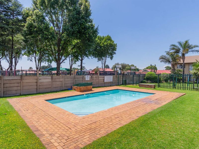 A well-maintained communal swimming pool area featuring brick paving, a surrounding lawn, and secure perimeter fencing within the apartment complex grounds.