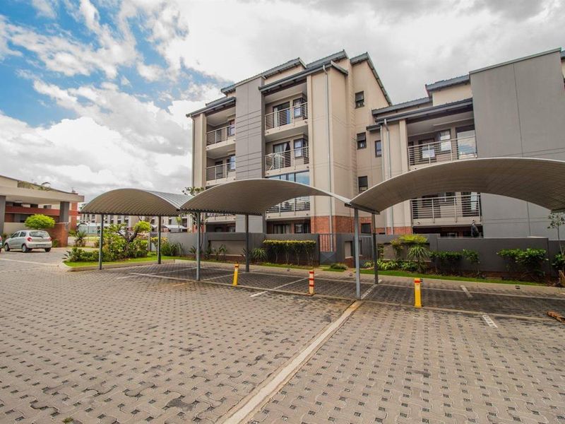 Exterior view of a modern apartment complex featuring covered parking bays, paved driveway, and multi-story residential units with balconies.