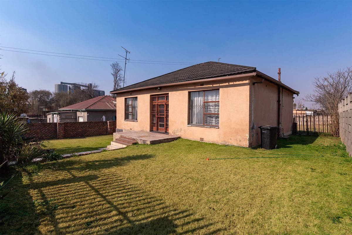Rear view of a detached house featuring a lawn, concrete patio, and security-fitted windows and doors, showing signs of exterior wear.