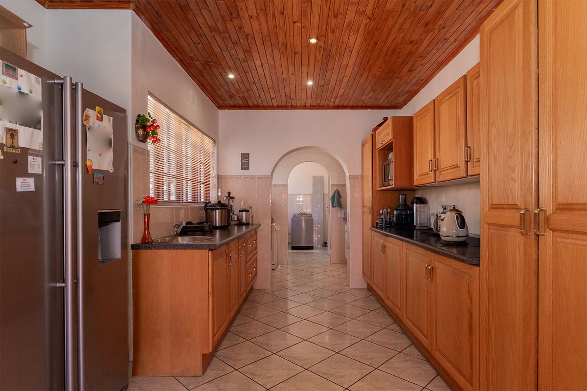 A functional galley-style kitchen featuring timber ceiling paneling, tiled flooring, ample laminate cabinetry, and stone countertops, leading through an archway to a utility area.