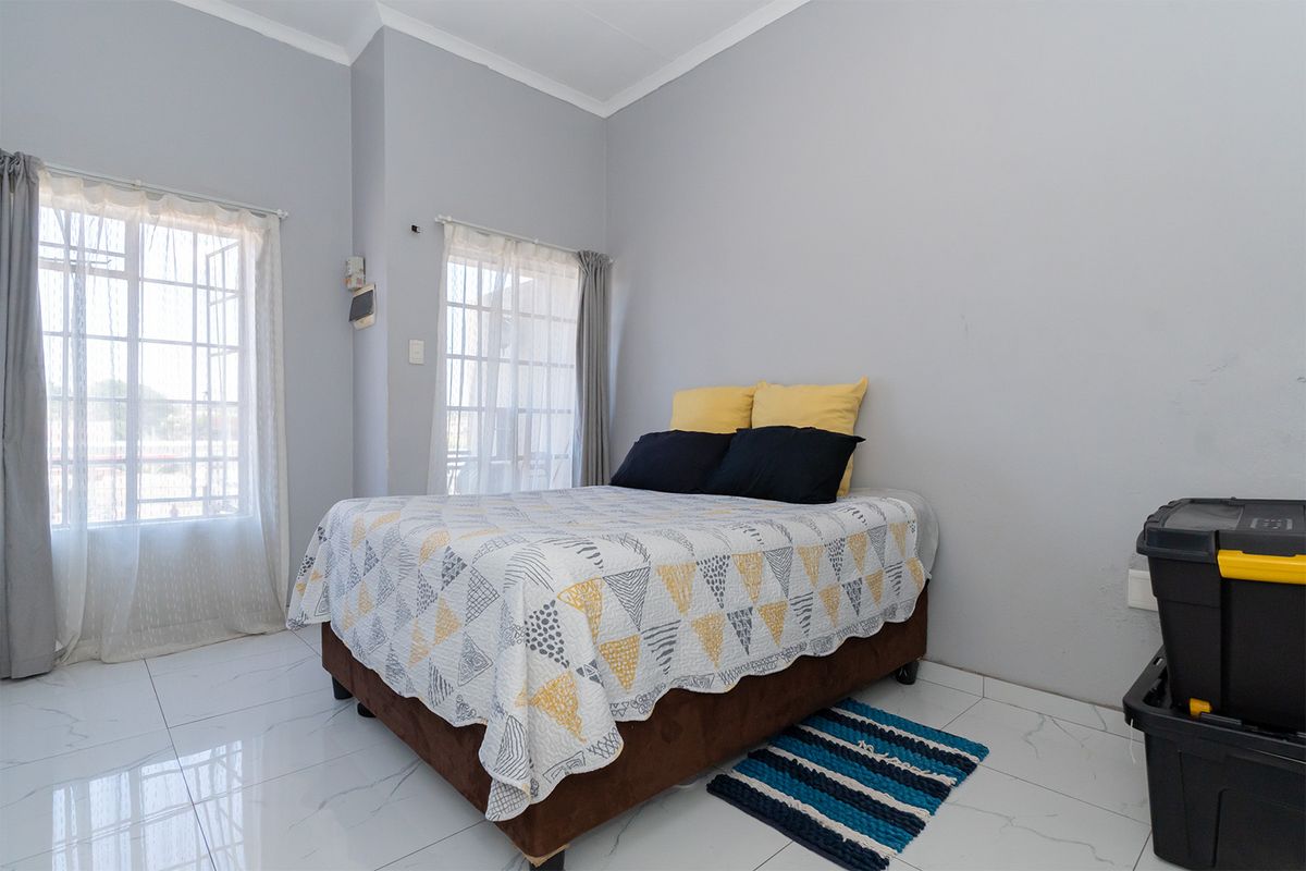 A bedroom featuring white tiled flooring, large windows with security bars, and neutral wall tones, providing a bright and functional living space.