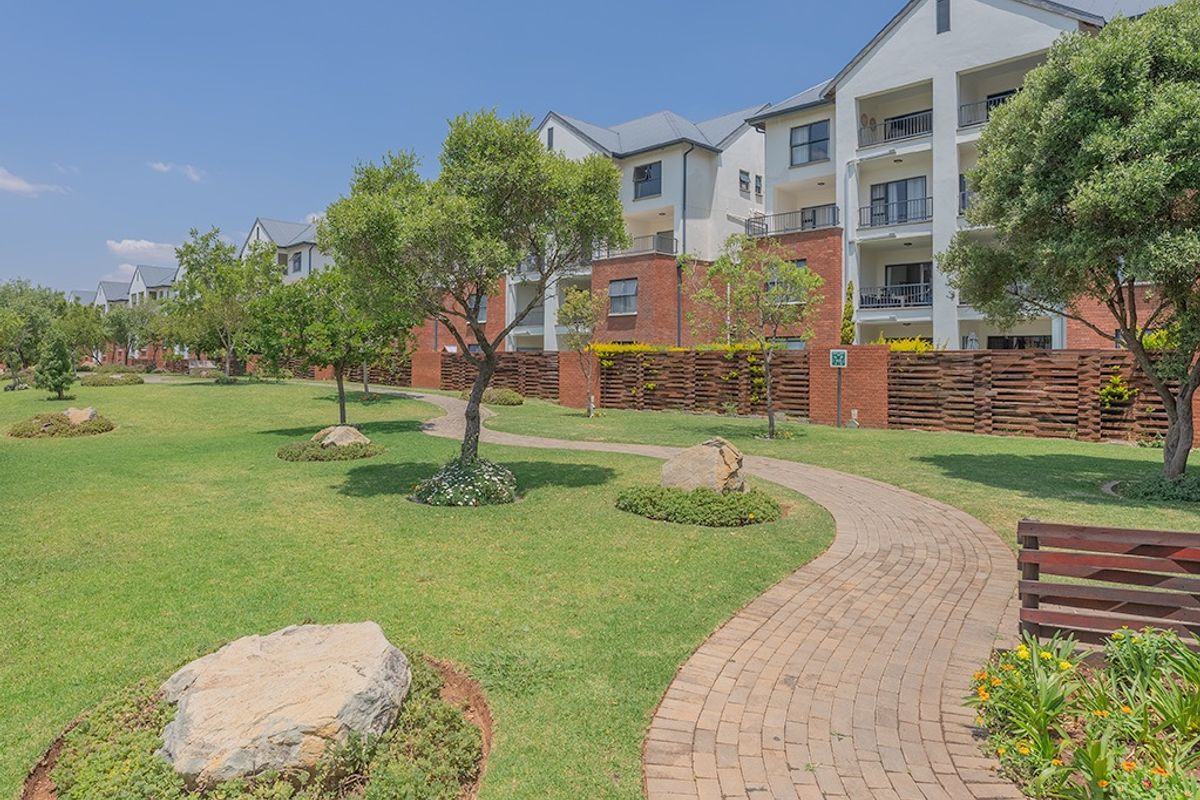 A well-maintained communal garden area featuring a paved walkway, manicured lawns, and mature trees, set against a multi-story apartment complex.