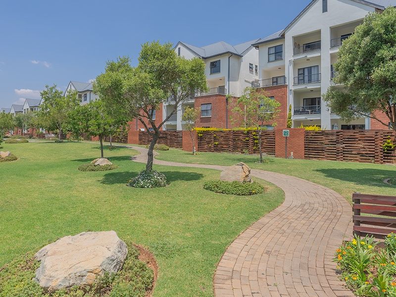A well-maintained communal garden area featuring a paved walkway, manicured lawns, and mature trees, set against a multi-story apartment complex.